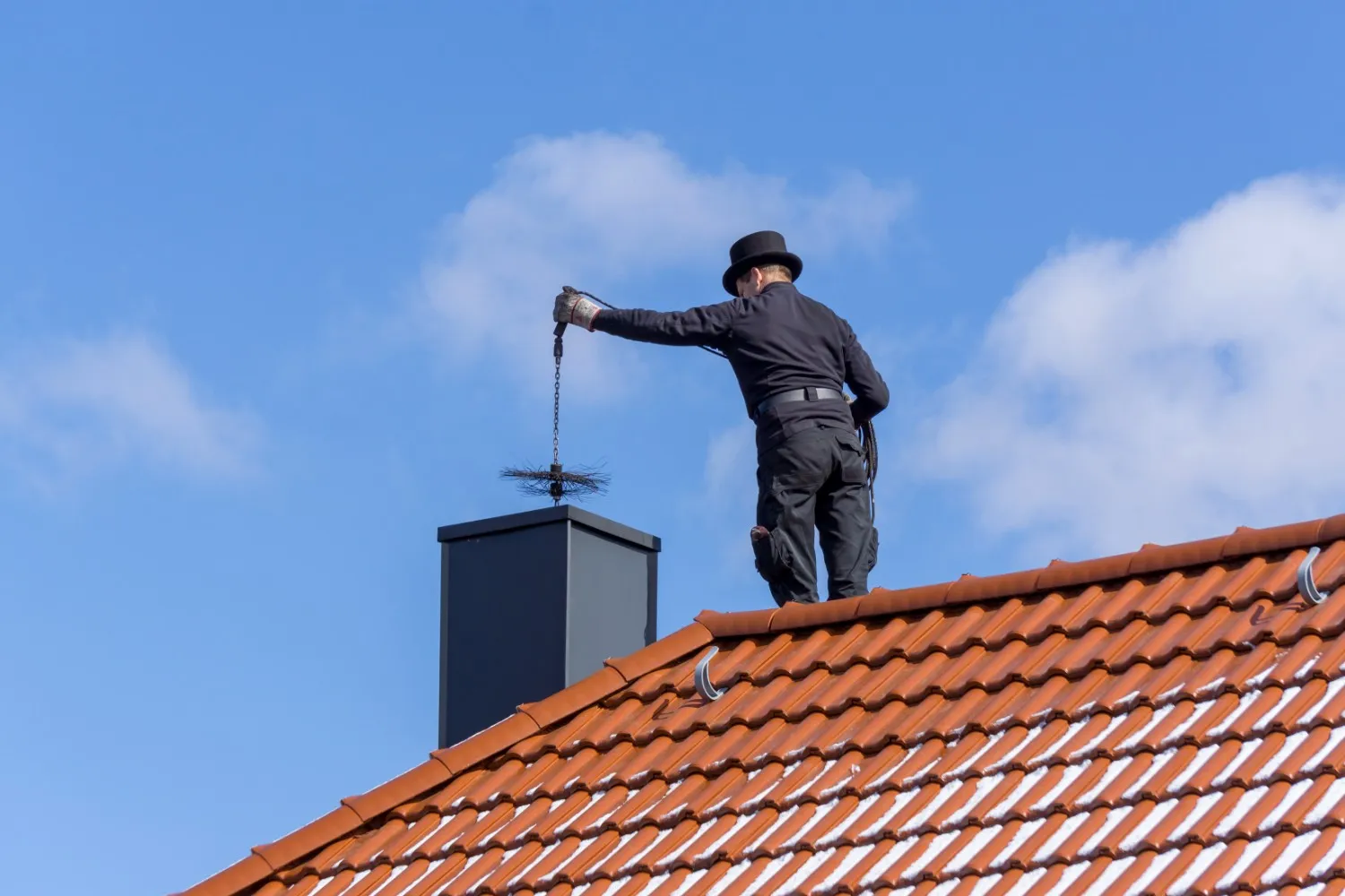 A chimney sweep in black clothing and a top hat standing on a tiled roof and cleaning a chimney.