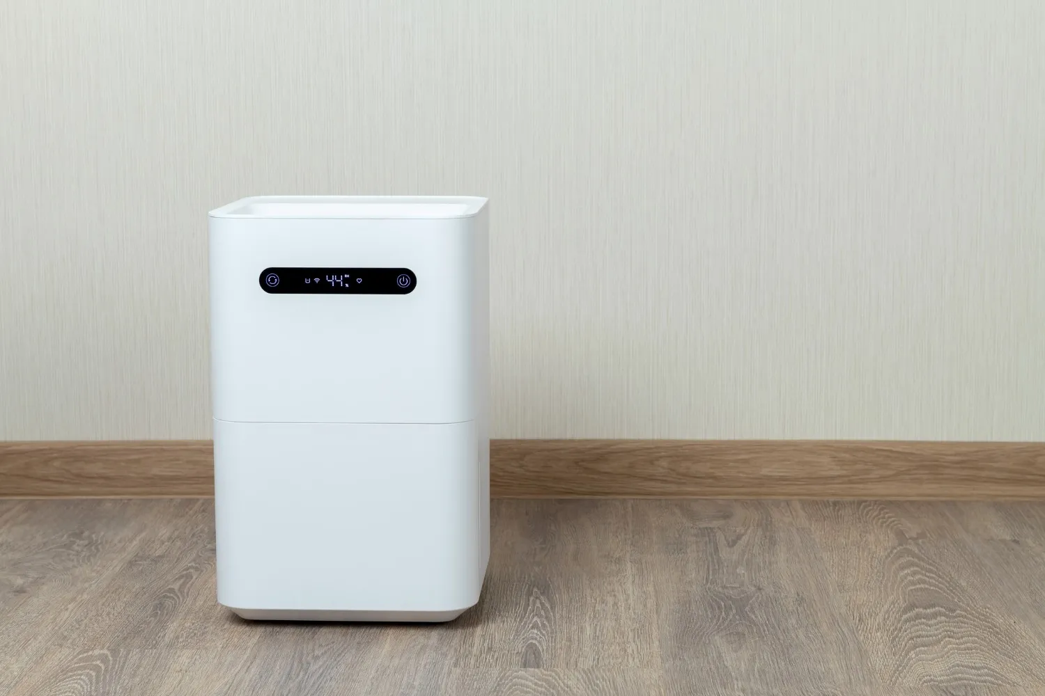 A square, white humidifier with a digital display showing 44% humidity sits on a wooden floor against a beige wall.