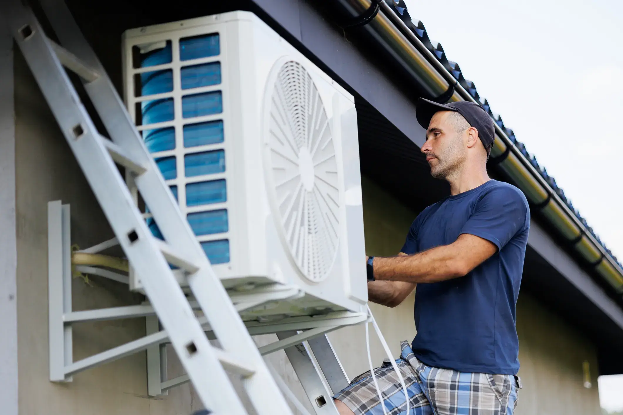 An HVAC technician on a ladder working on an outdoor air conditioning unit attached to a house.
