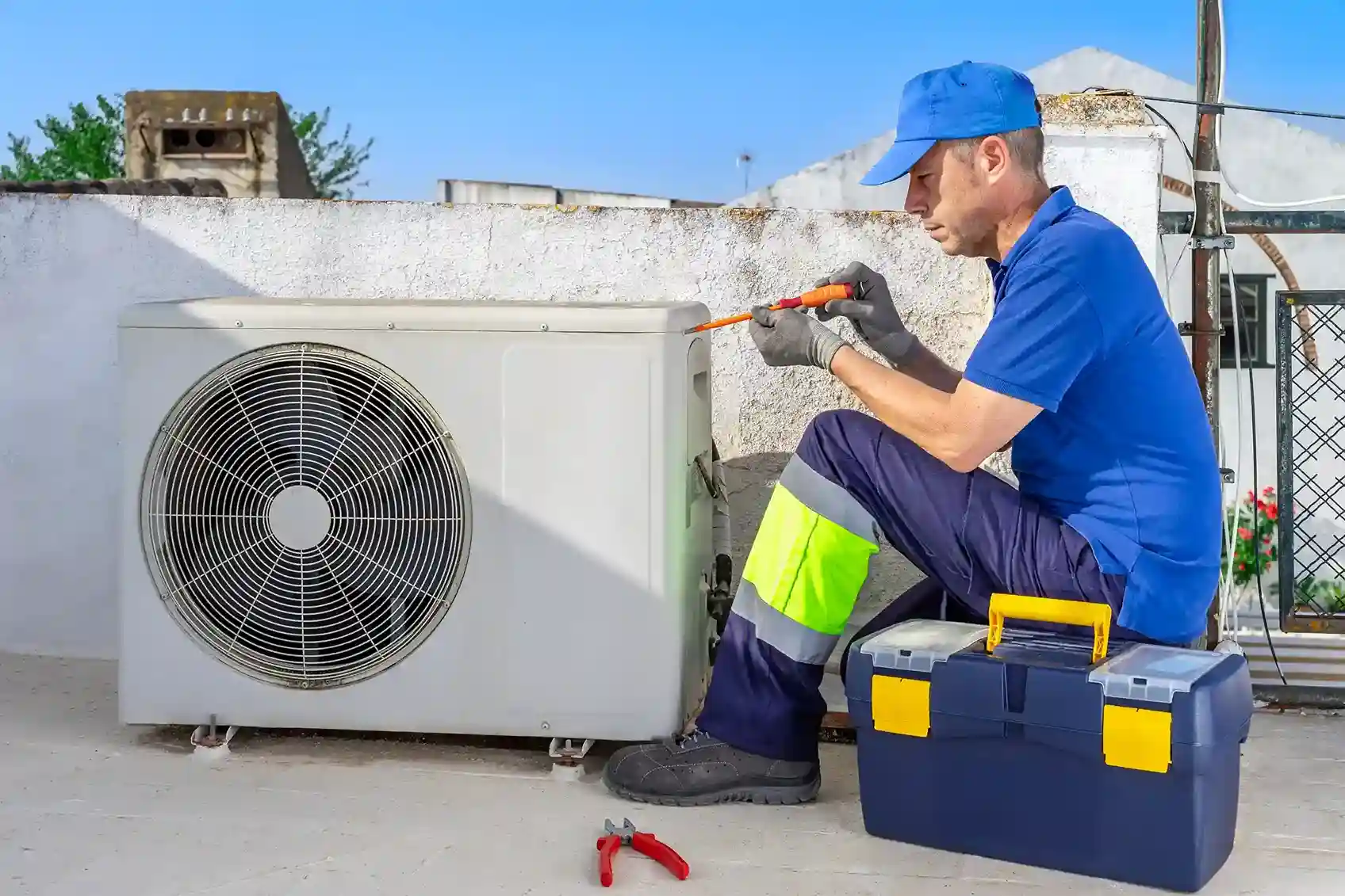 An HVAC technician in a blue uniform and cap repairing an outdoor air conditioning unit on a rooftop.