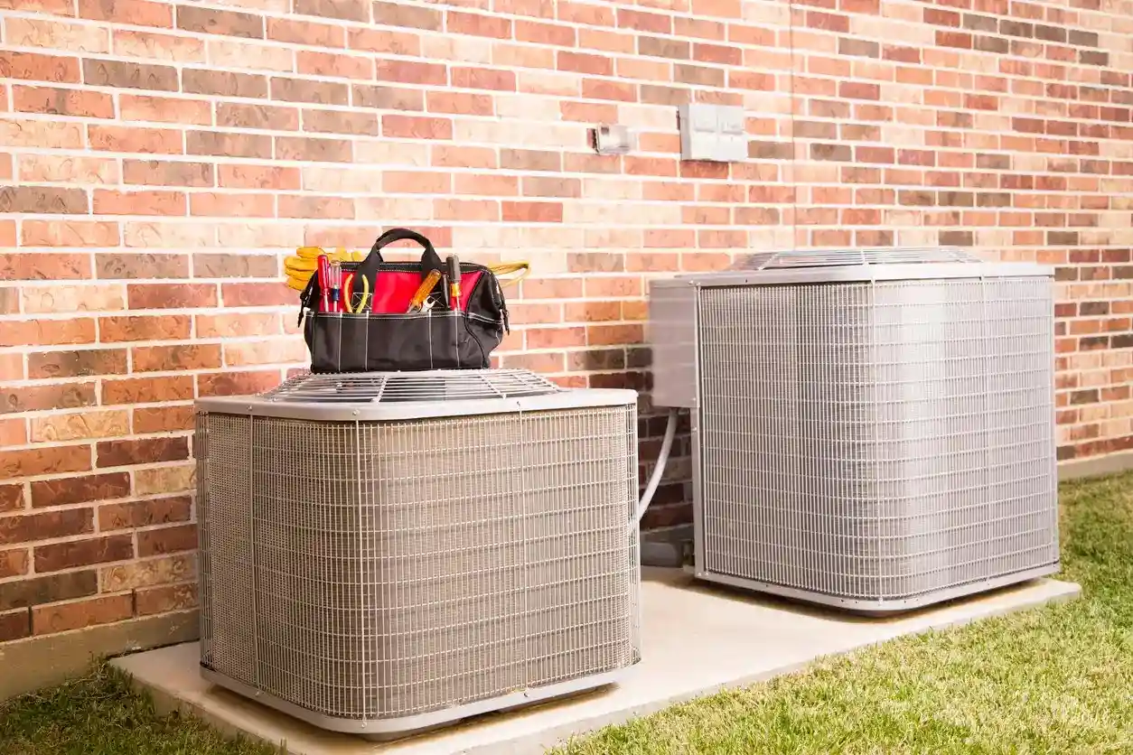 Two new outdoor HVAC condenser units next to a brick wall with a tool bag sitting on one.