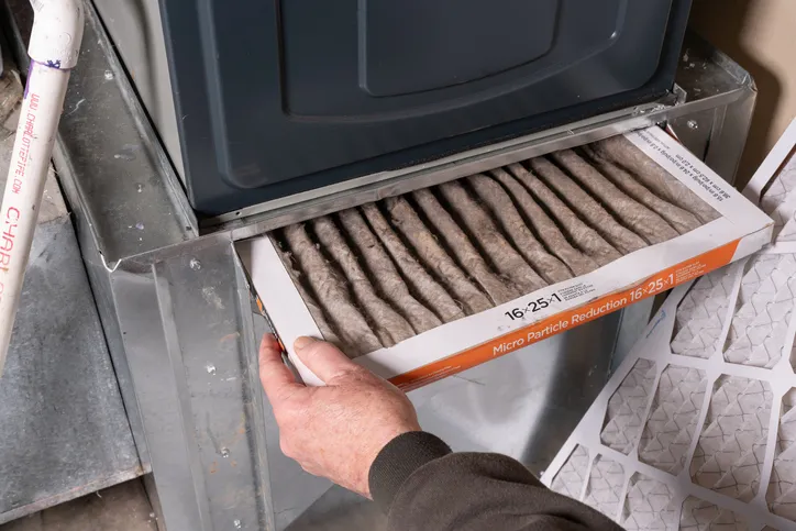 A hand is pulling a very dirty, dark brown air filter out of the return air duct slot of a furnace, indicating a need for repair or replacement service.