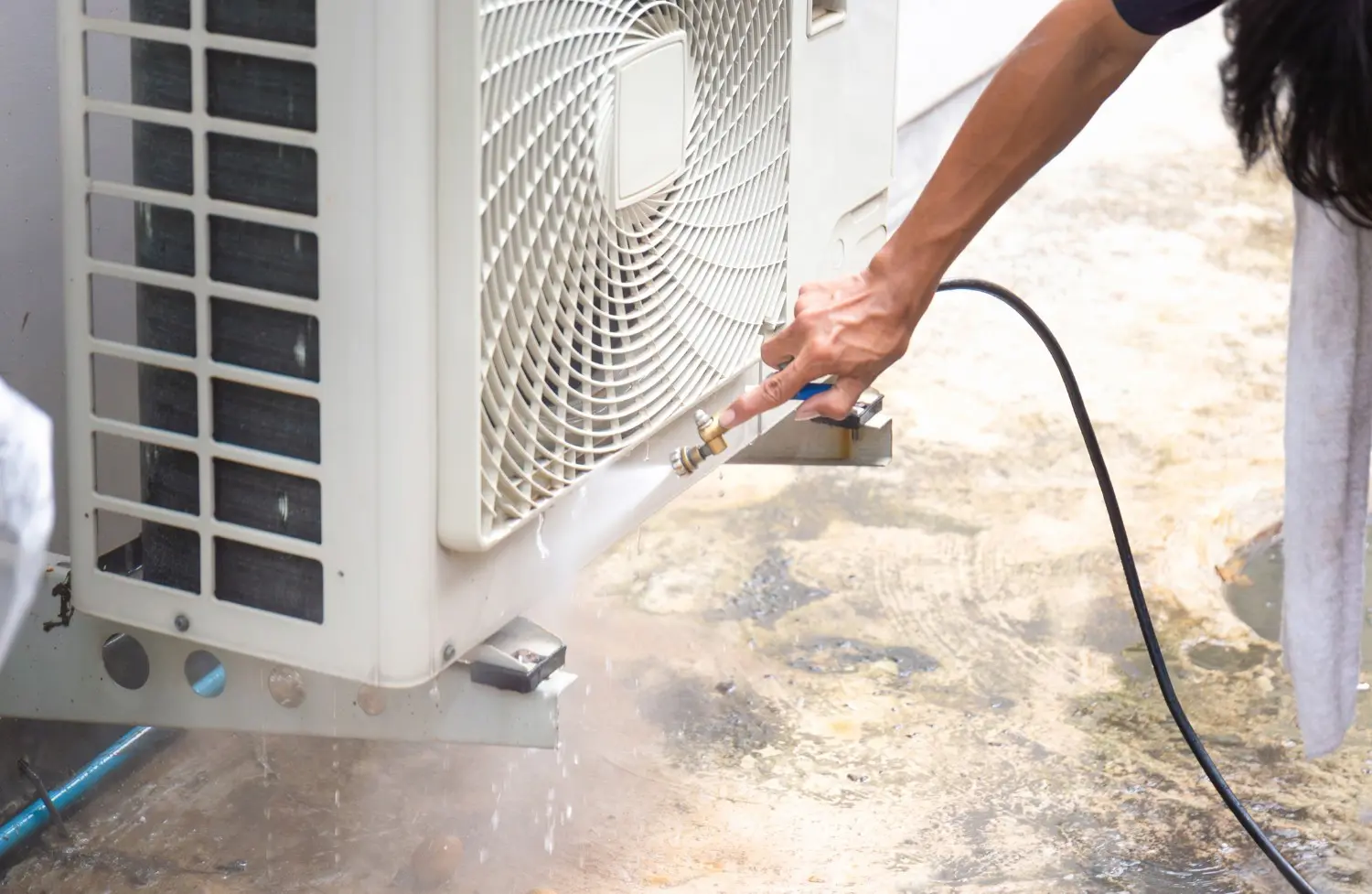 A close-up shot of a person's arm using a black hose or sprayer to clean the fins and fittings on the side of an outdoor air conditioning unit, creating a splash of water and dirt on the concrete floor.
