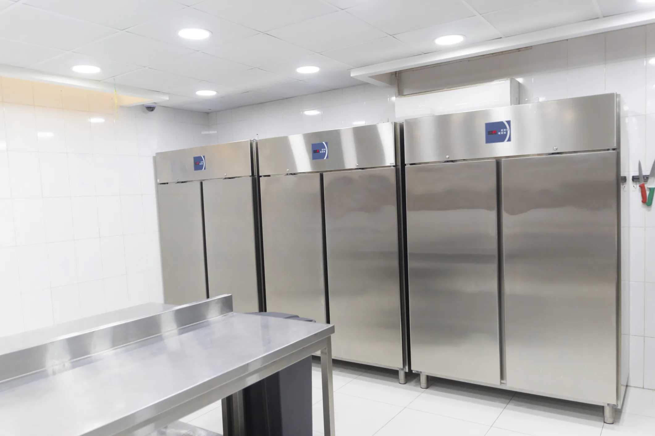 A row of four large, stainless steel, double-door commercial refrigerators/freezers stands against a white tiled wall in a professional kitchen or food preparation area. A stainless steel work table is visible in the foreground.