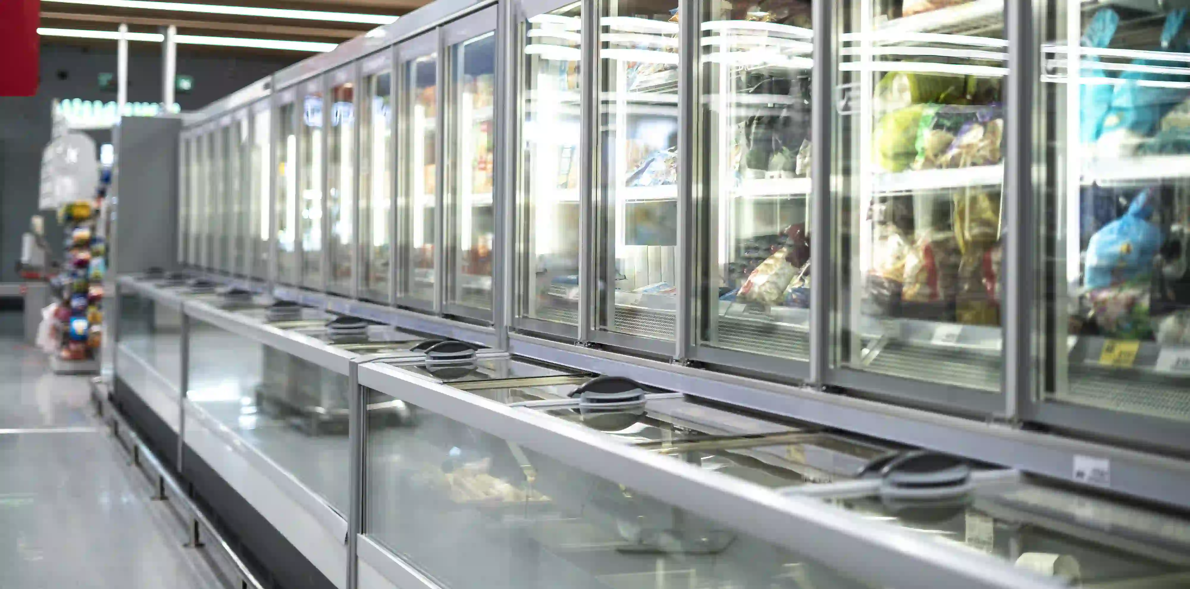 A long aisle in a supermarket or grocery store featuring a continuous line of refrigerated and frozen display cases. The upper section consists of upright glass-door freezers stocked with packaged food, while the lower section is an open-top refrigerated display case with horizontal glass lids.