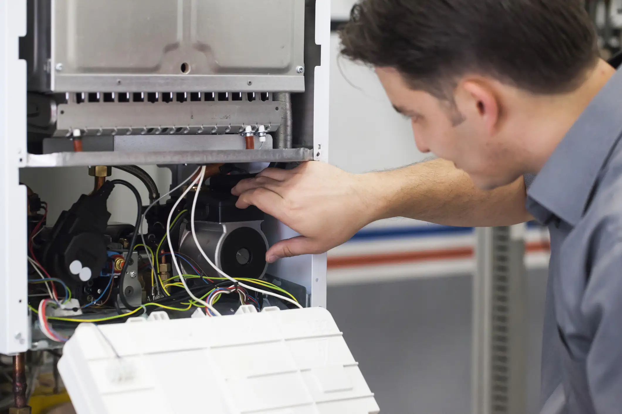 A male HVAC technician in a gray shirt is leaning over an open white boiler or modern furnace unit, with his hand reaching toward the internal components, including wiring and a motor, while holding the access panel open with his other hand.