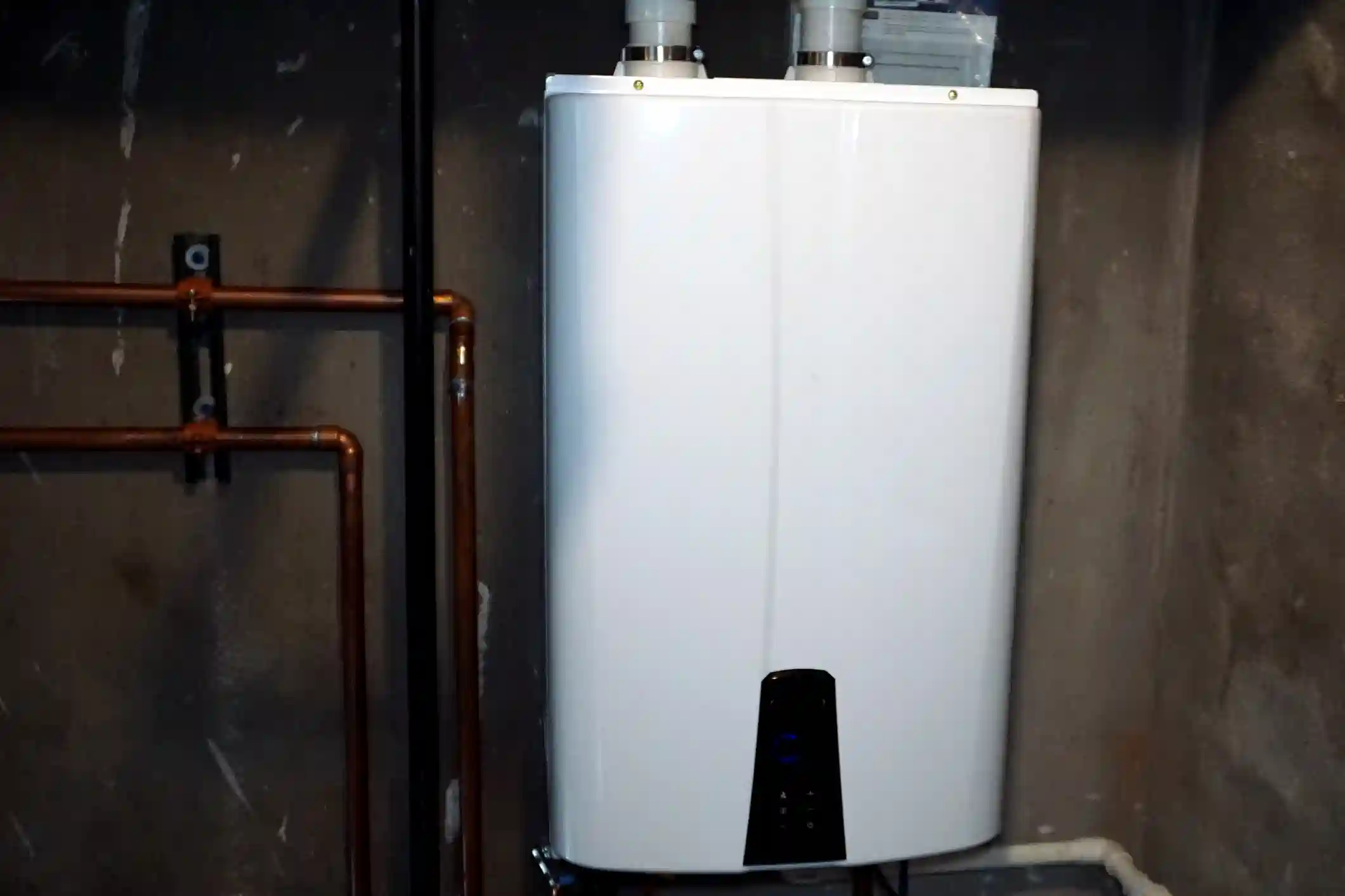 A new, white electric tankless water heater is mounted on a dark, unfinished basement wall. To the left, copper plumbing lines are visible, mounted to the wall with black brackets. The water heater has a black control panel at the bottom.