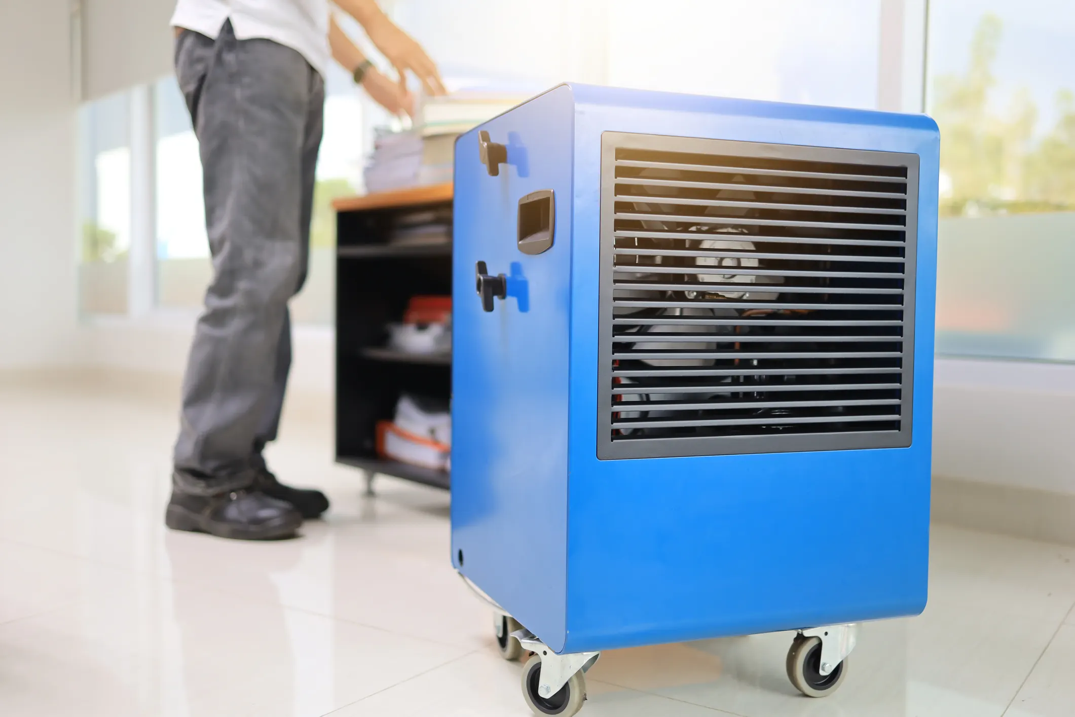 A large, blue commercial-style dehumidifier on wheels is positioned in the foreground of a bright, modern office space with large windows. In the background, a person wearing a white shirt and dark jeans is visible from the waist down, organizing items on a dark shelf.