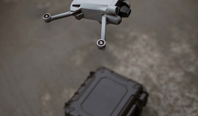 Compact quadcopter drone hovers above rugged protective carrying case on concrete
