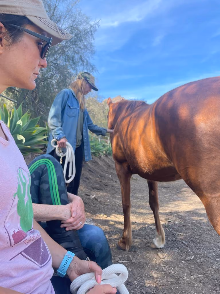 Two women outdoors with hats near a brown horse on a dirt path under a blue sky.