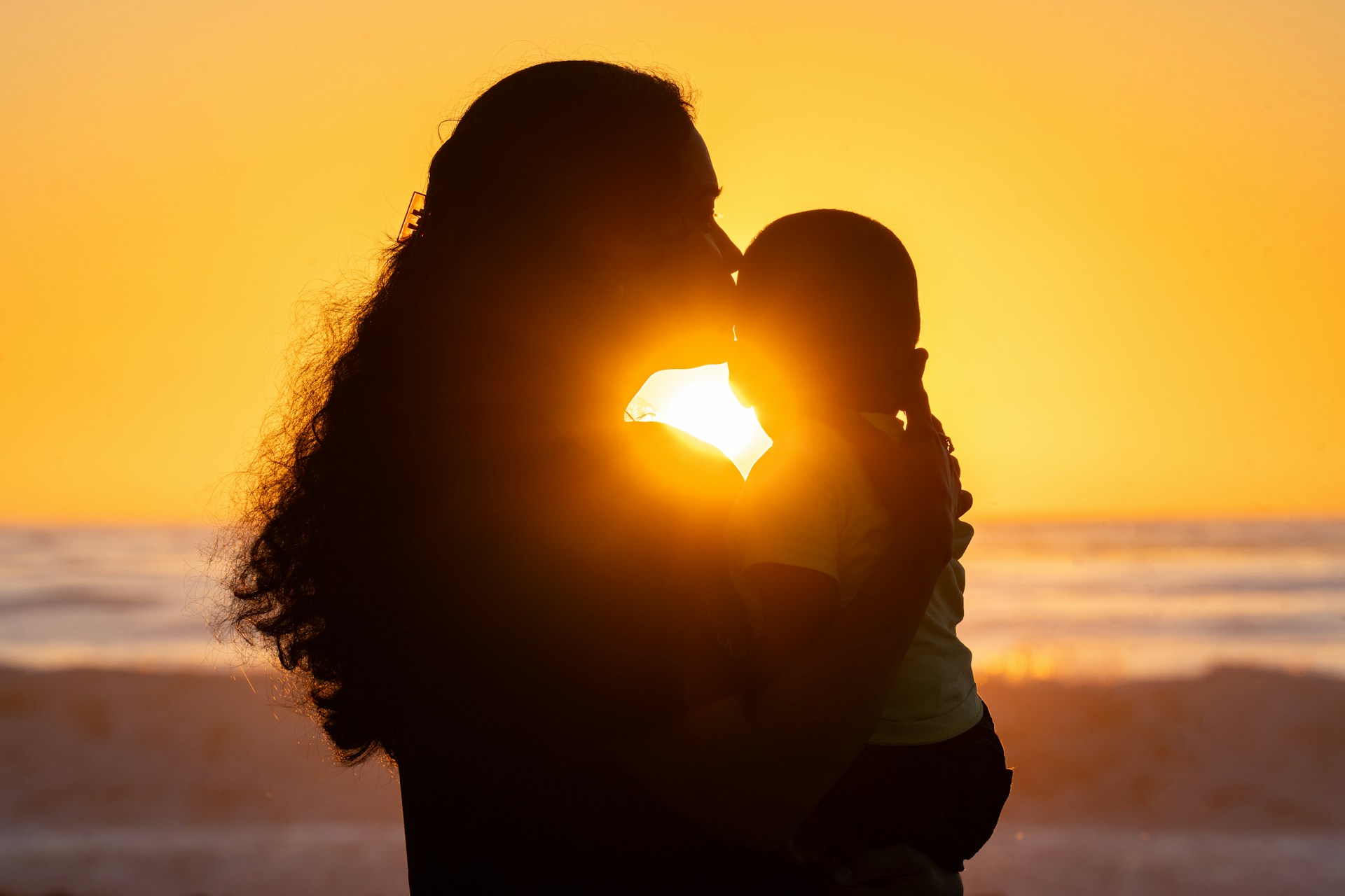 Silueta de una mujer abrazando a un niño contra un atardecer dorado en el mar.