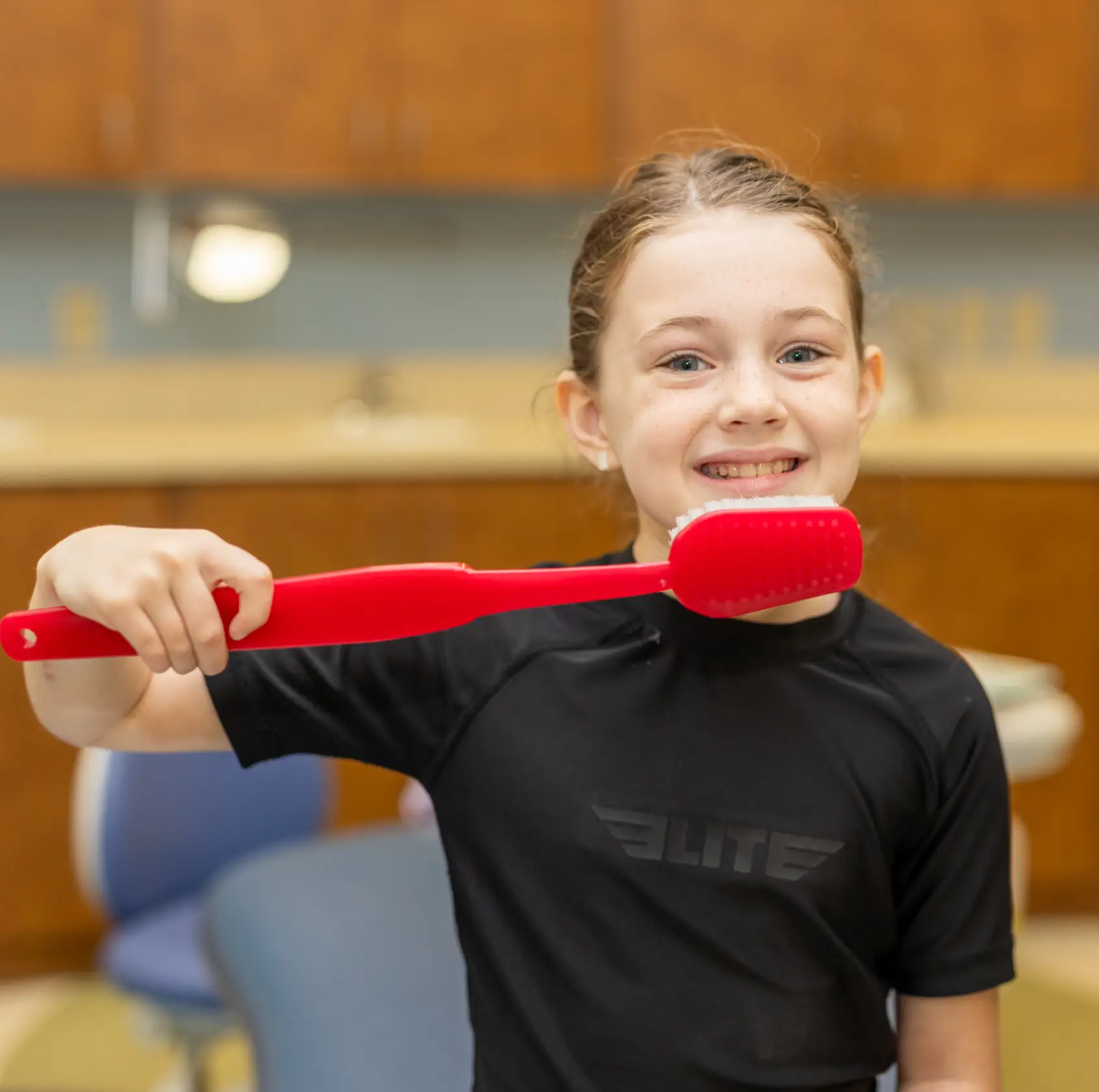 Smiling girl holding a large red toothbrush in a dental office setting.