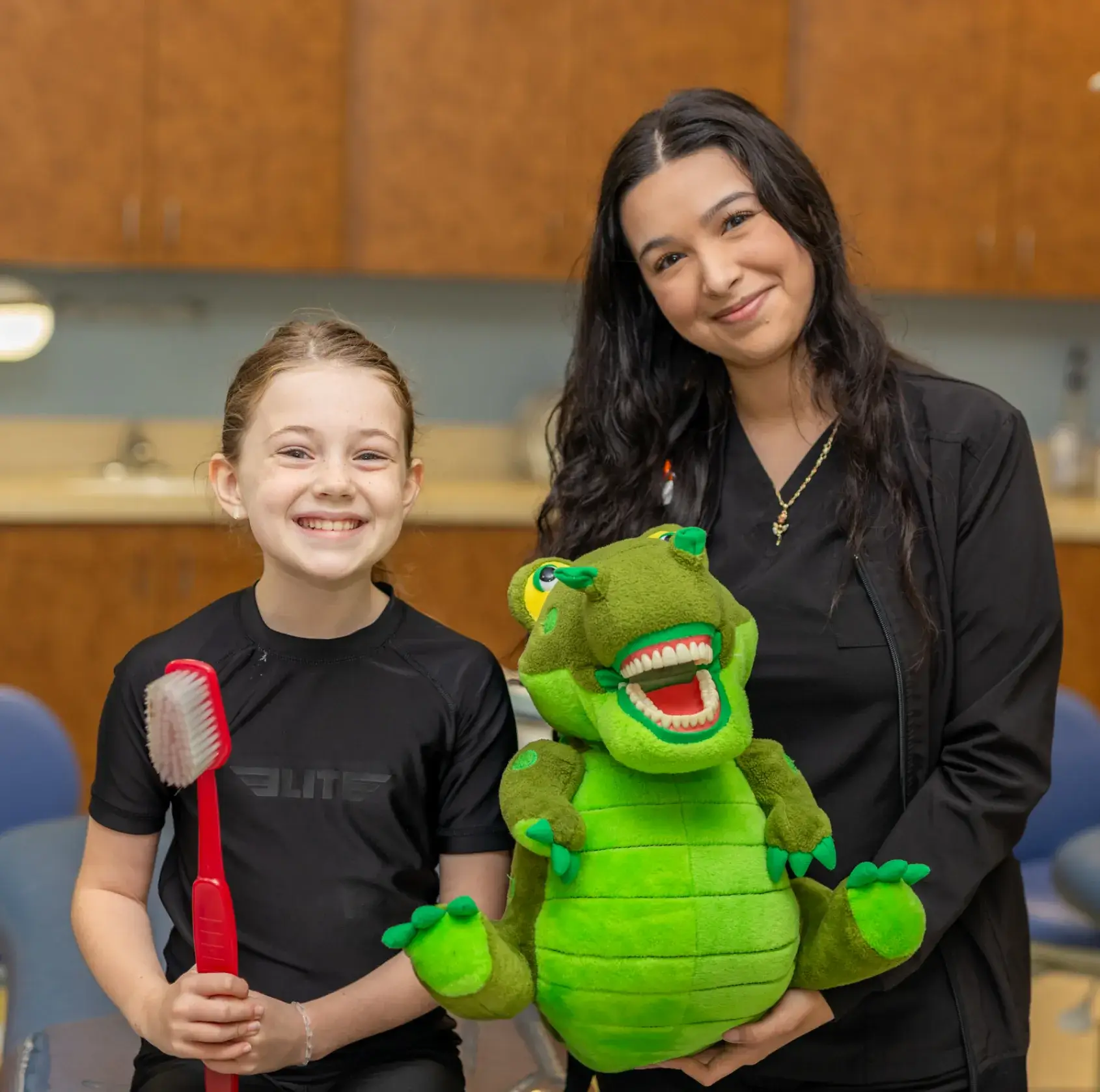 A young girl holds a large toothbrush, standing next to a woman holding a green plush dinosaur.