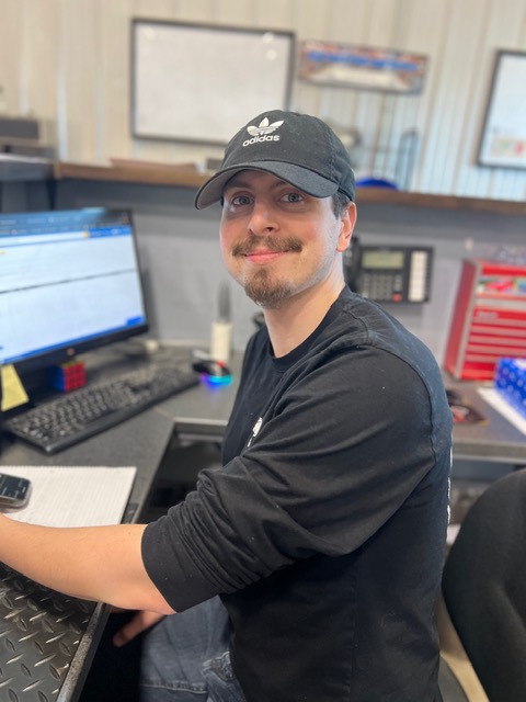 Smiling man wearing a black Adidas cap and sweatshirt sitting at a desk with a computer monitor.