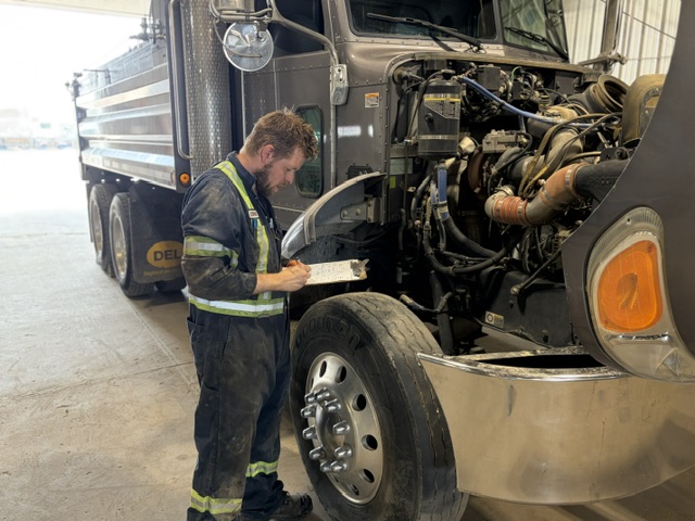Man in workwear performing SGI safety inspection and taking notes on clipboard next to a large truck with open engine compartment.