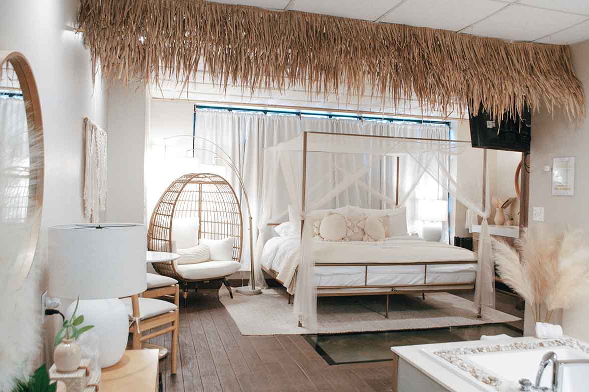 A beach-inspired bedroom with a white canopy bed, a thatched ceiling accent, and a unique glass floor panel over a glowing water feature, enhancing the island ambiance.