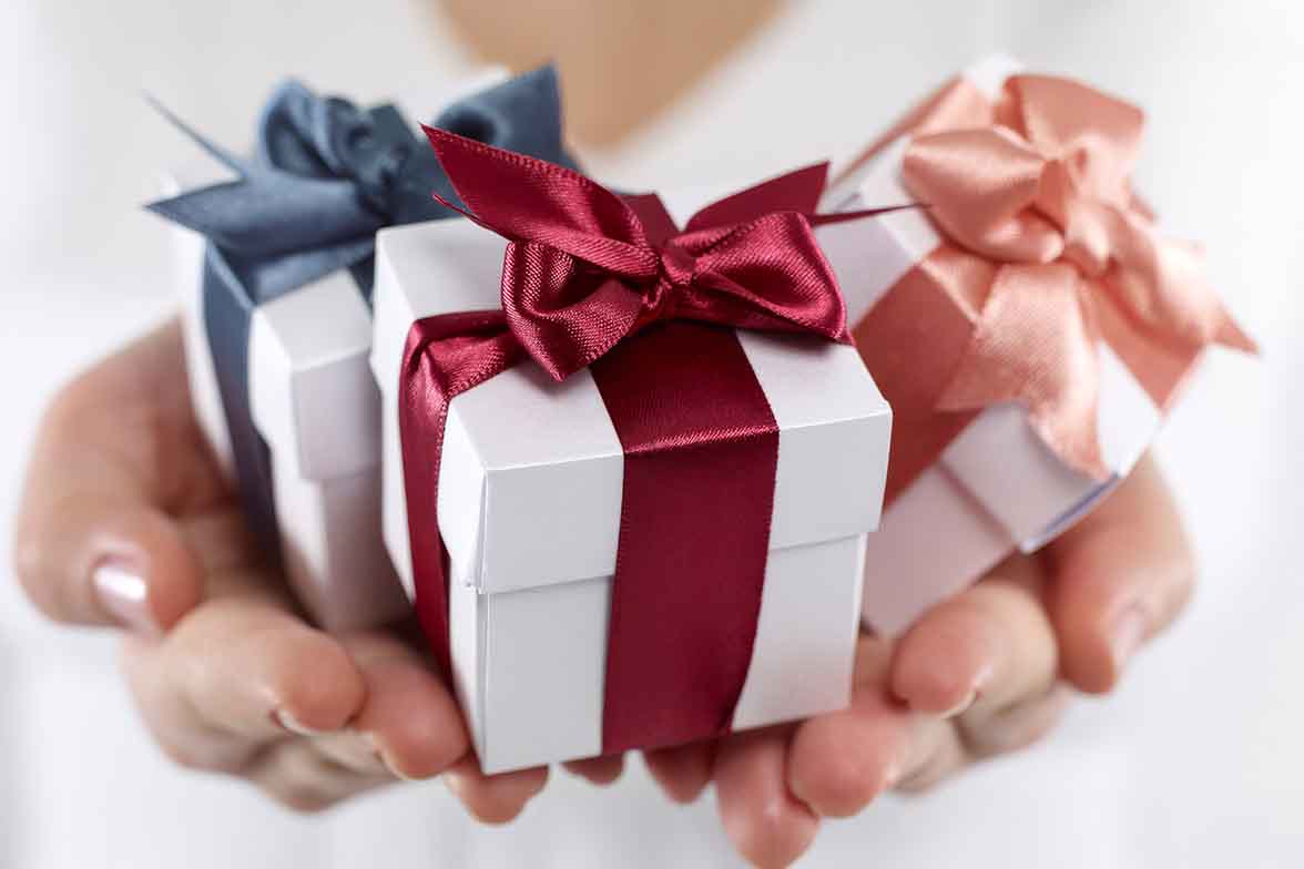 Hands holding three small white gift boxes with red, blue, and peach satin ribbon bows.