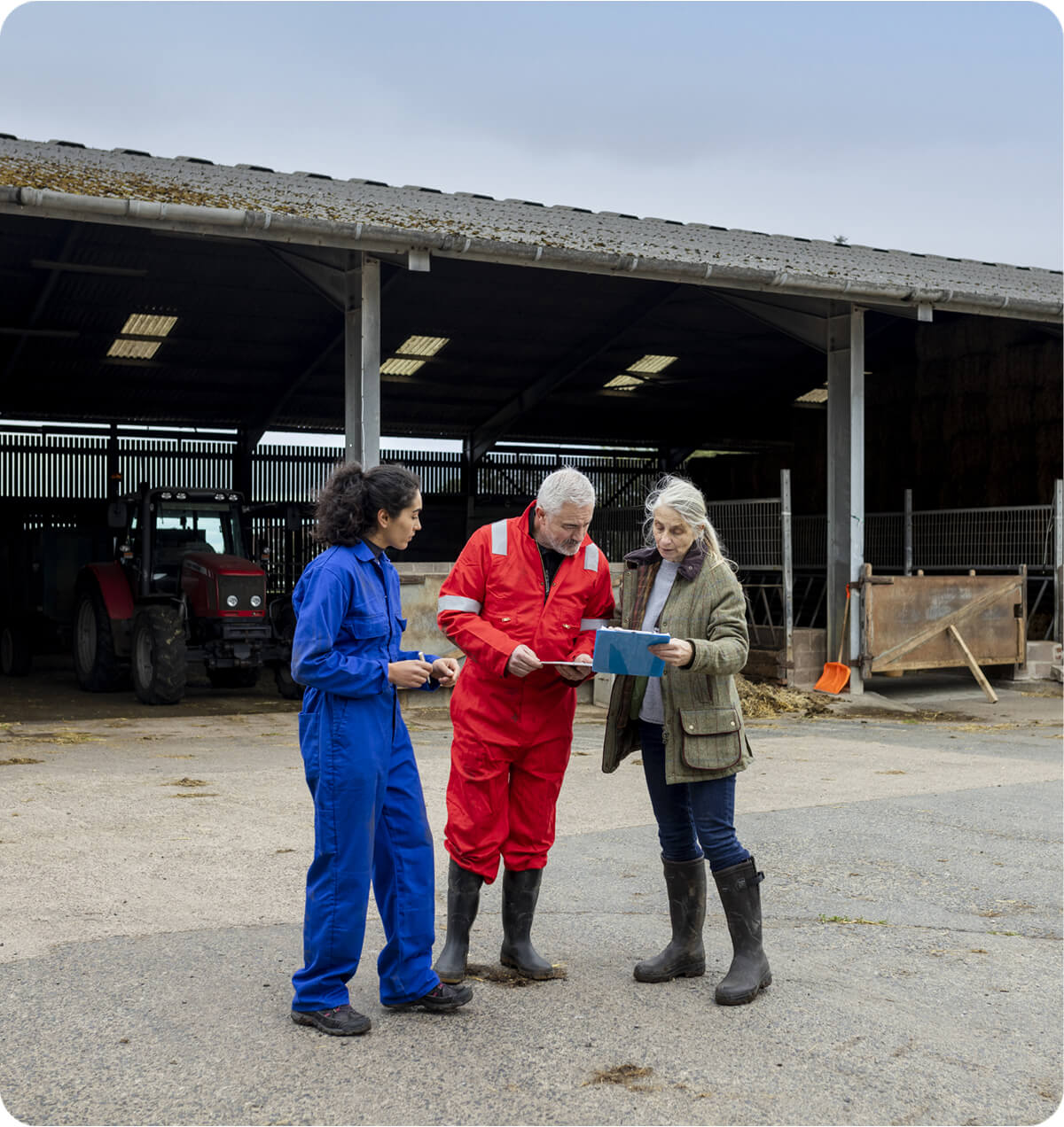 A man and a women in overalls, standing next to another women who's showing them something on a clip board. There is a farm shed in the background.