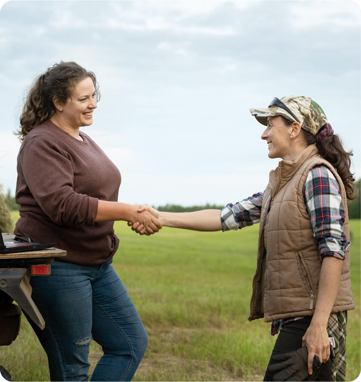 Two women standing in a paddock, leaning against a truck's deck with a laptop, shaking hands.