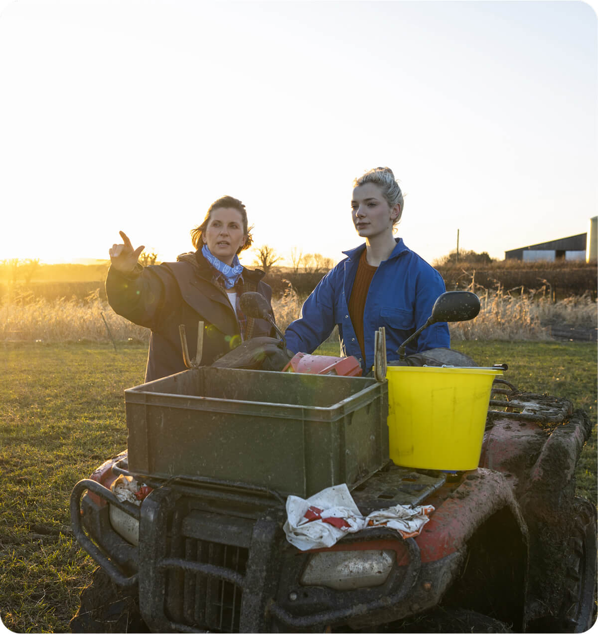 A women sitting on a muddy quad motorbike next to a tractor, with another women standing beside her pointing and talking to her.