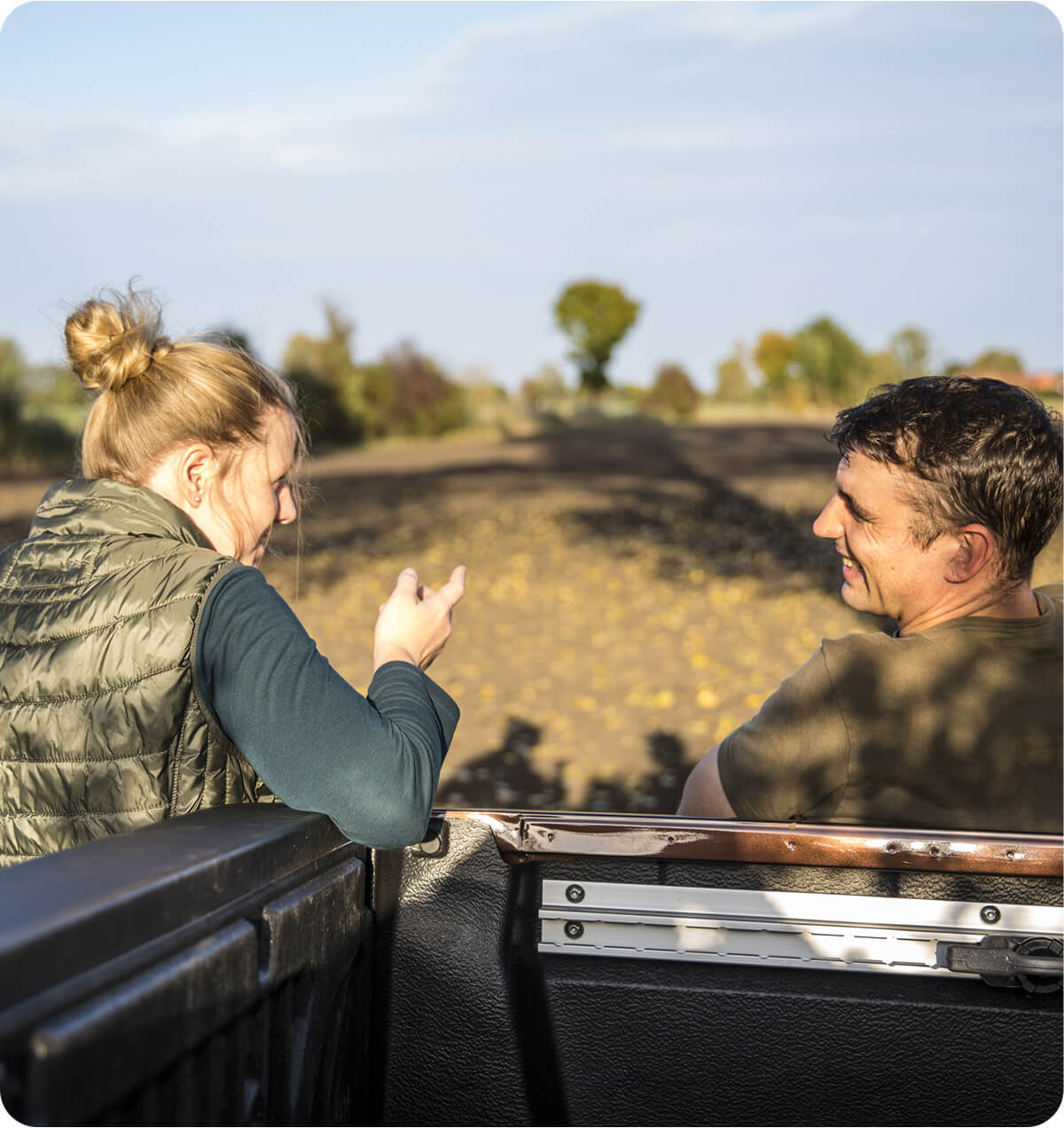 A women and a man leaning against the back of a truck in a cultivated paddock talking, with the women gesturing towards the crop.