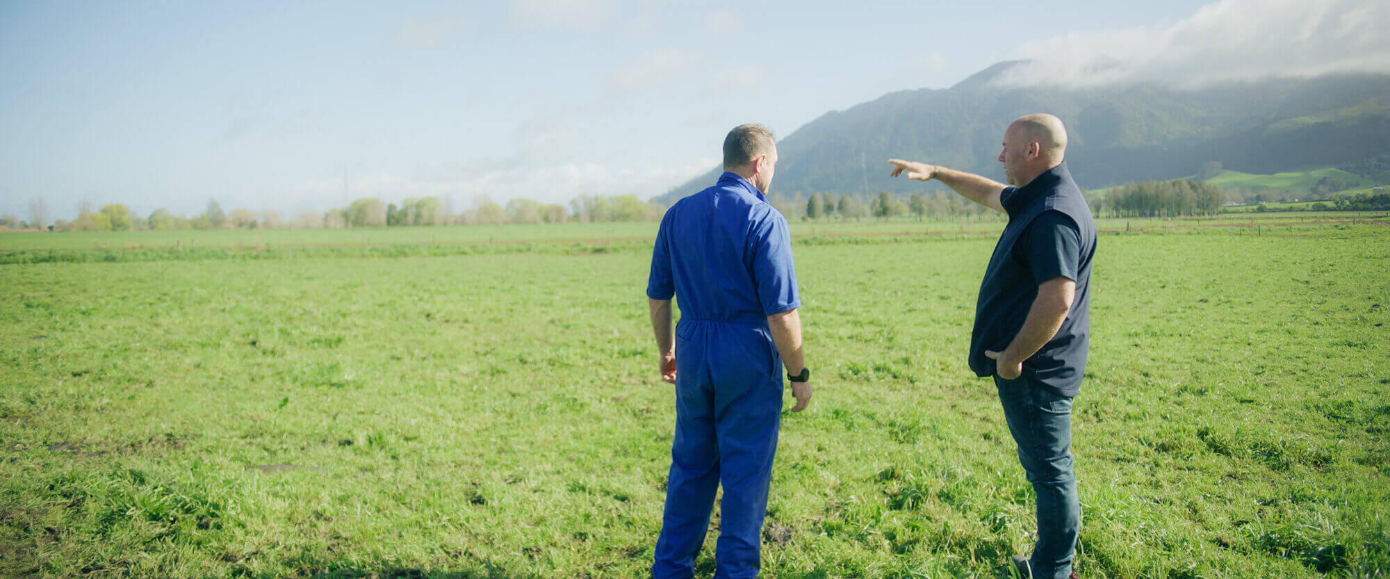 A farmer in overalls standing in a paddock with another man who is pointing off into the distance.