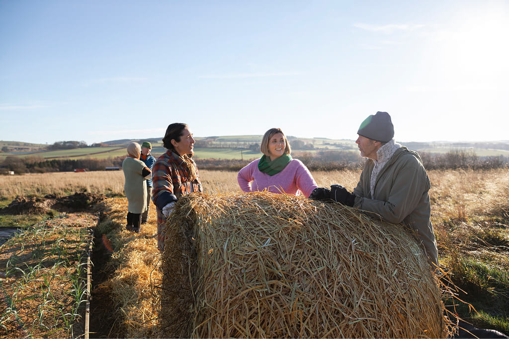 Two women and a man leaning against a hay bail in a paddock talking, with two people behind them talking.