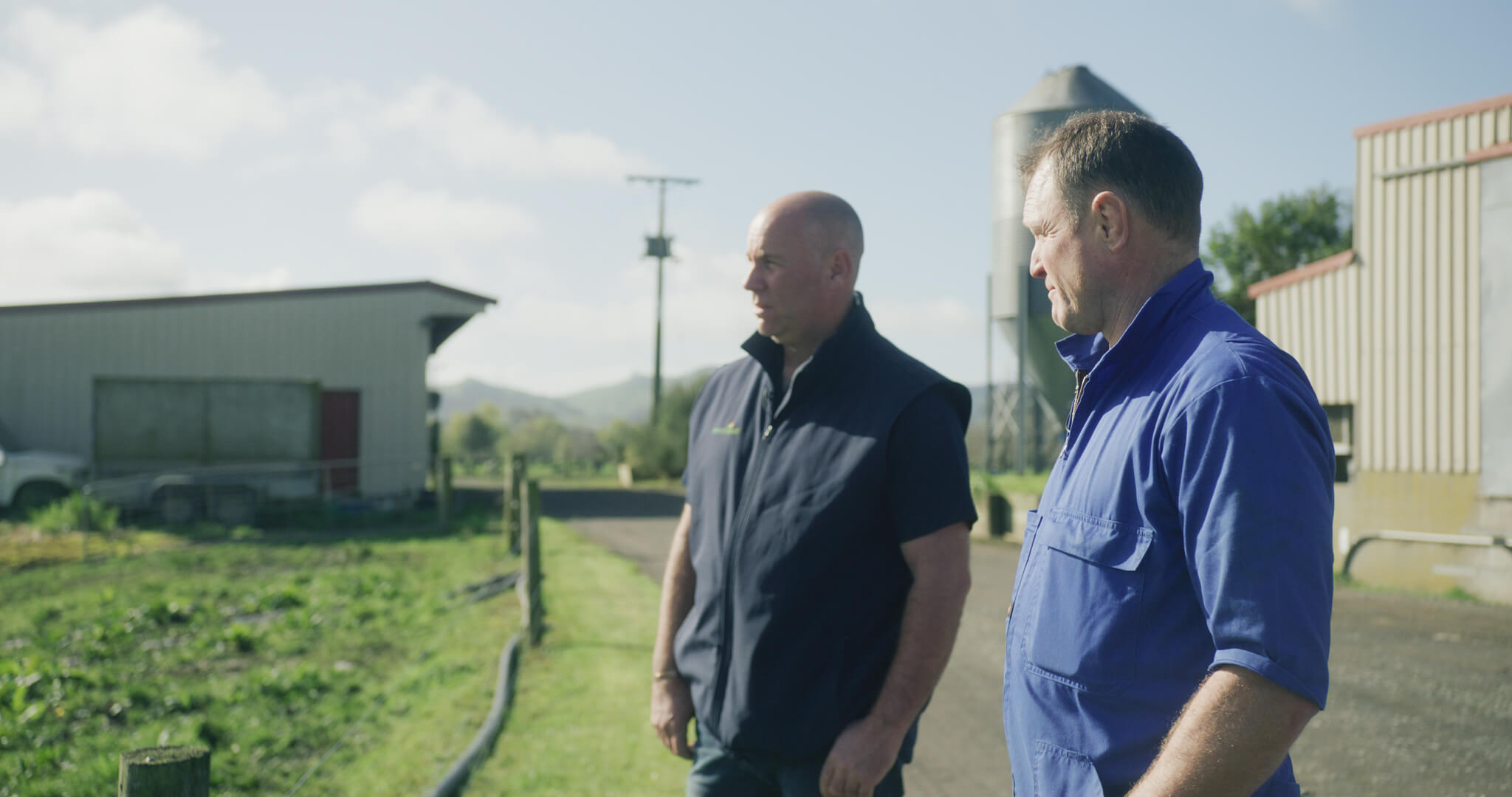 A farmer in overalls looking out into a farm yard with another man. 