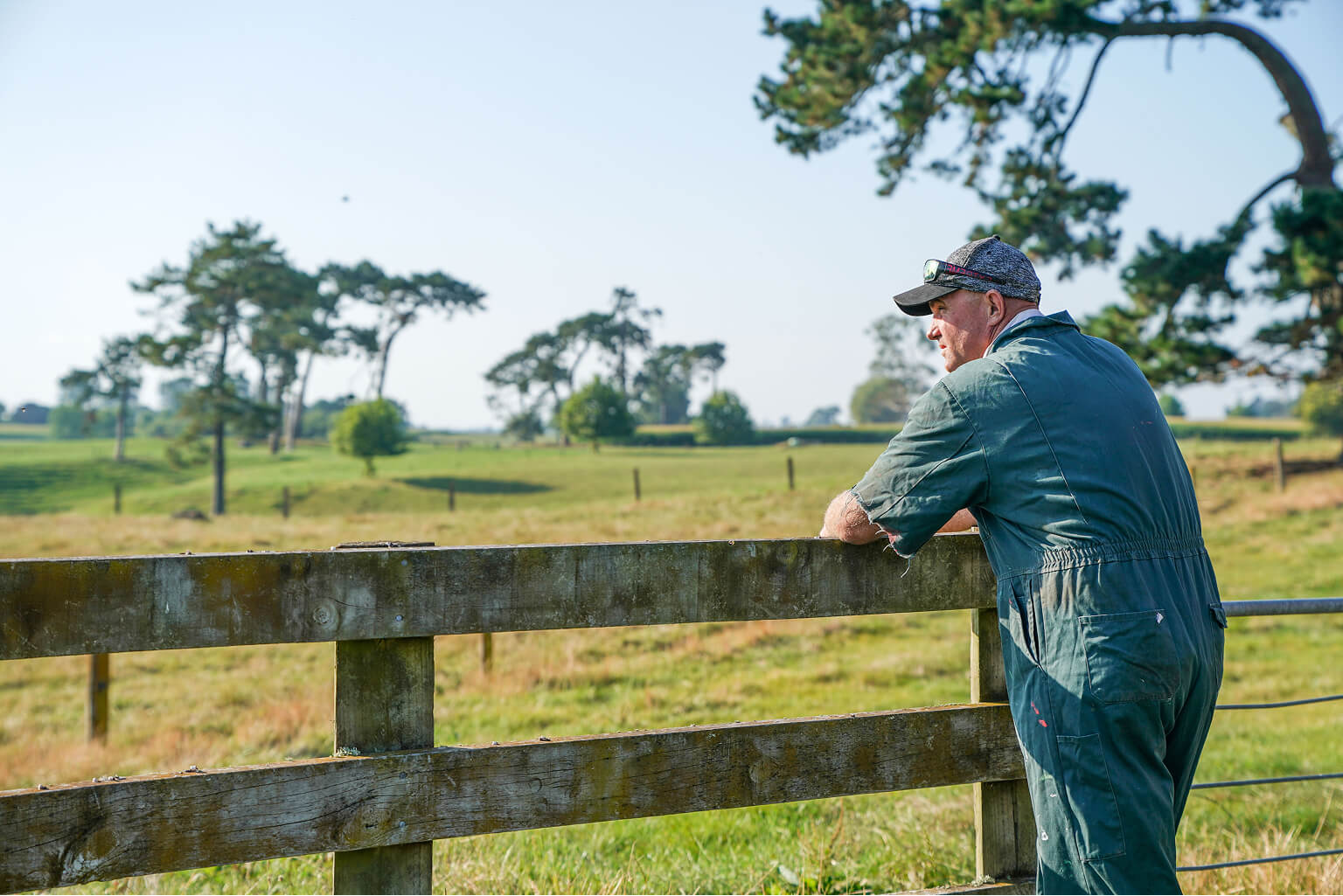 A farmer leaning against a fence looking out to a farm.