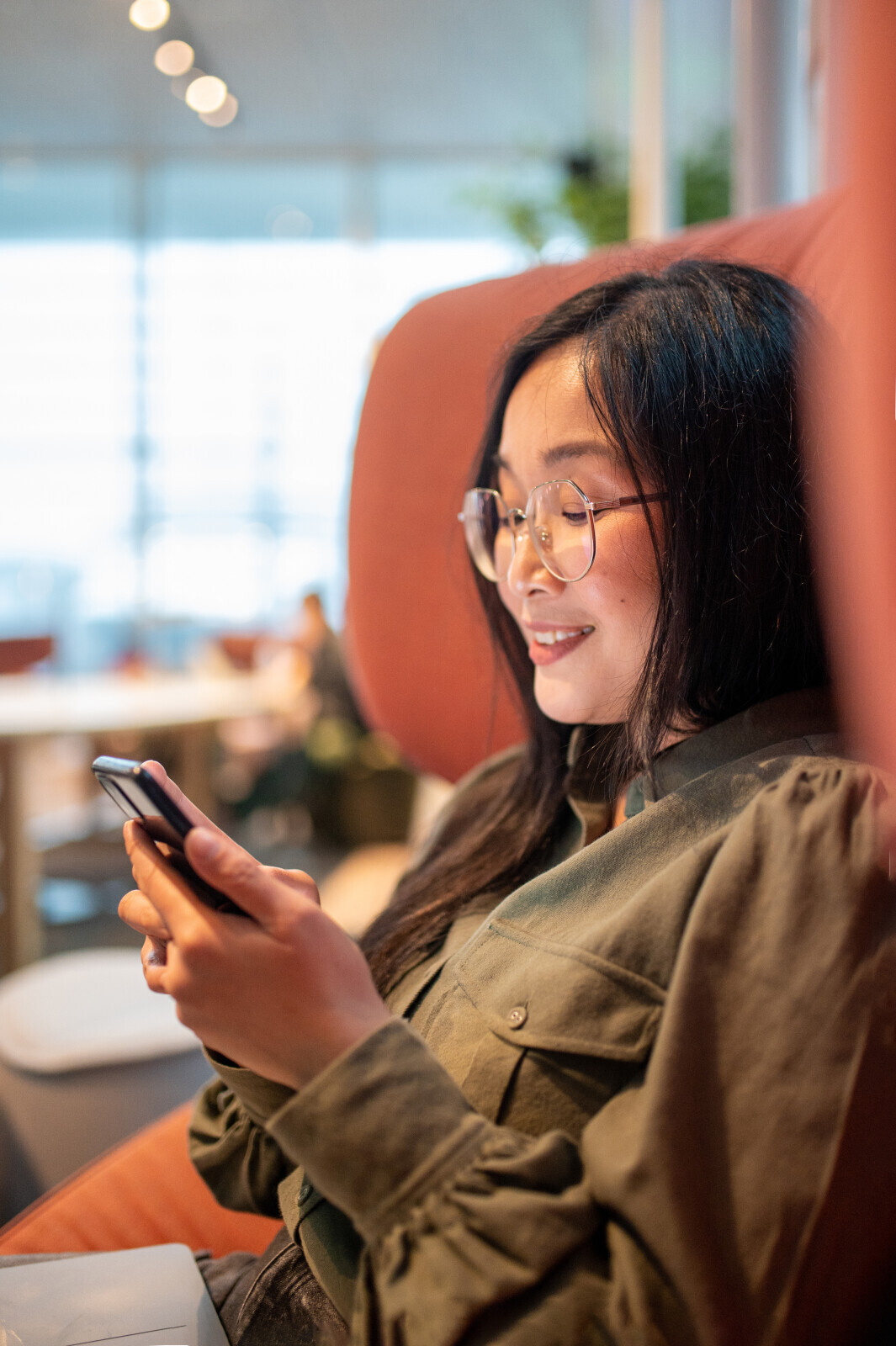 Woman with glasses smiling while looking at her smartphone sitting in a cozy indoor space.