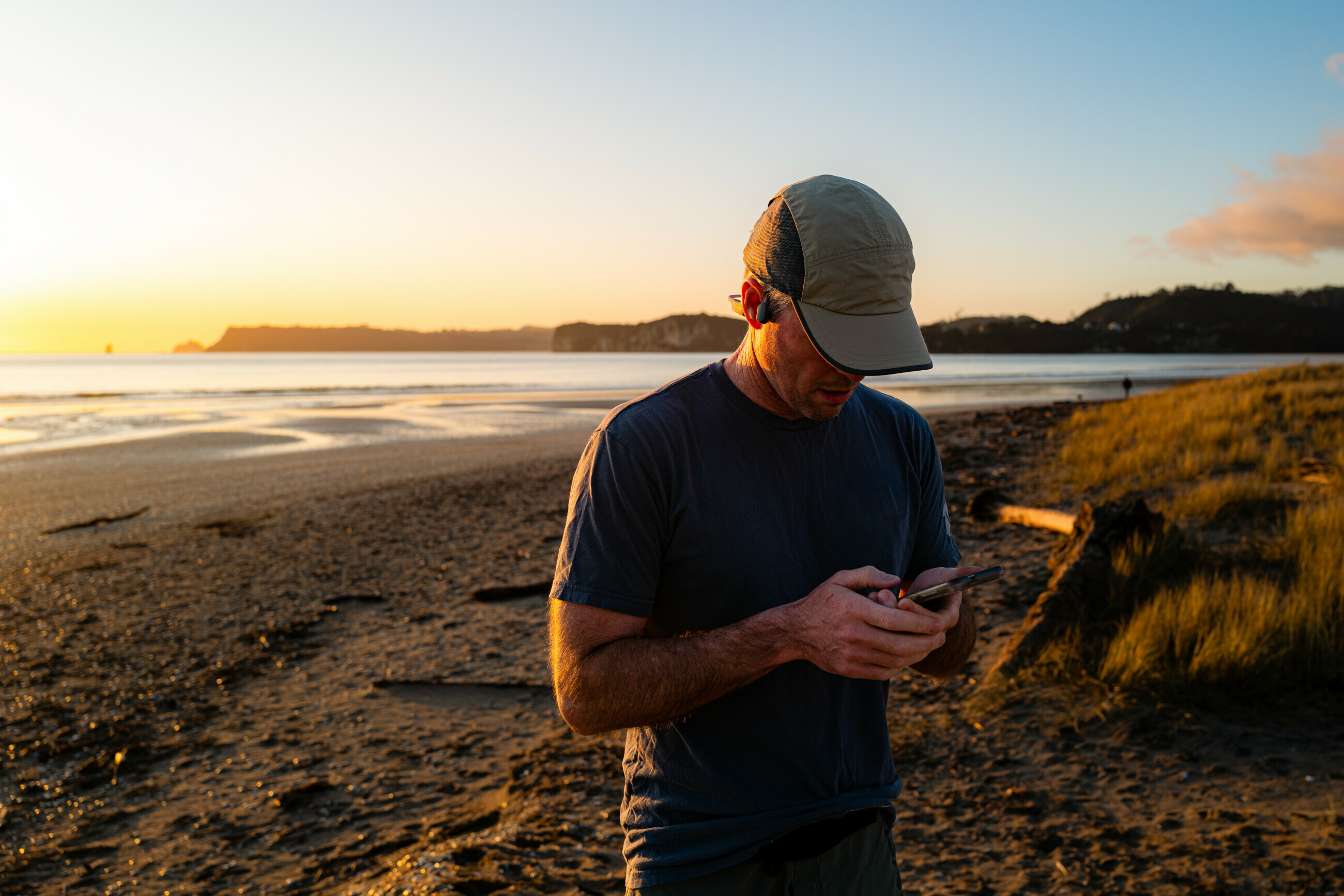 Man wearing cap and dark shirt looking at phone on a sandy beach during sunset.