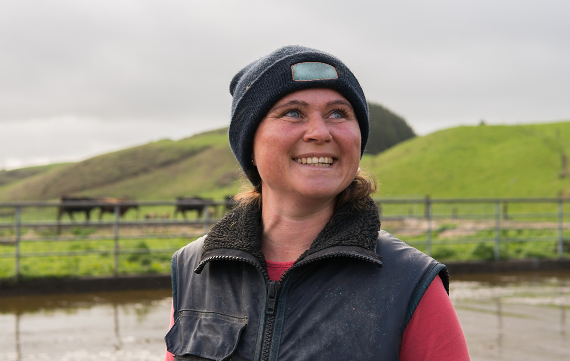 Smiling woman wearing a black beanie and vest stands outdoors by a fenced green pasture with cows.