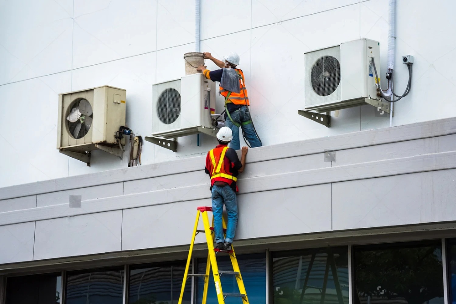 Workers installing air conditioning units on building exterior using ladder