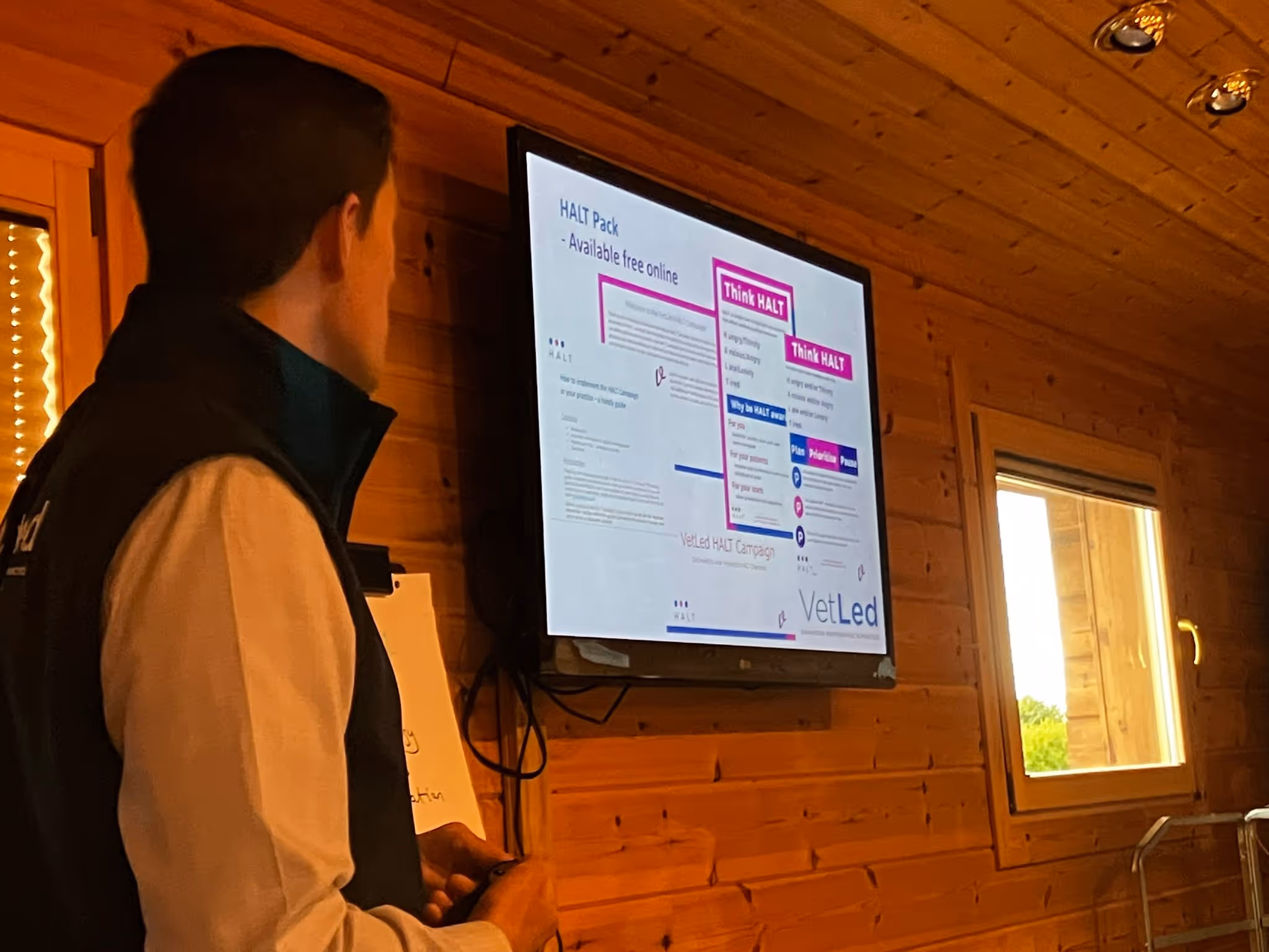 A man presents a slide about the "HALT Pack" from VetLed on a wall-mounted screen in a wooden room.