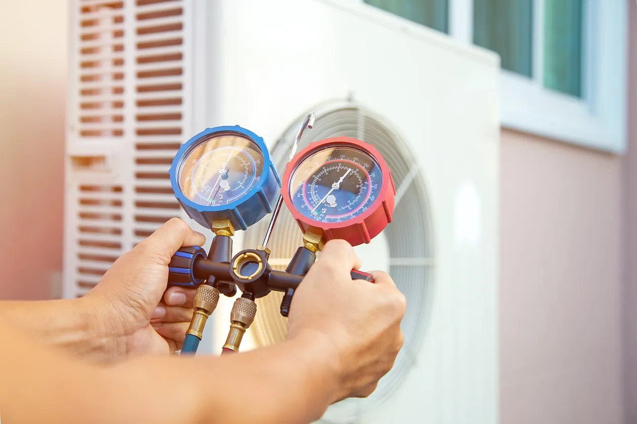 A close-up of a person's hands holding a manifold gauge set with red and blue dials, about to connect them to a heat pump unit.