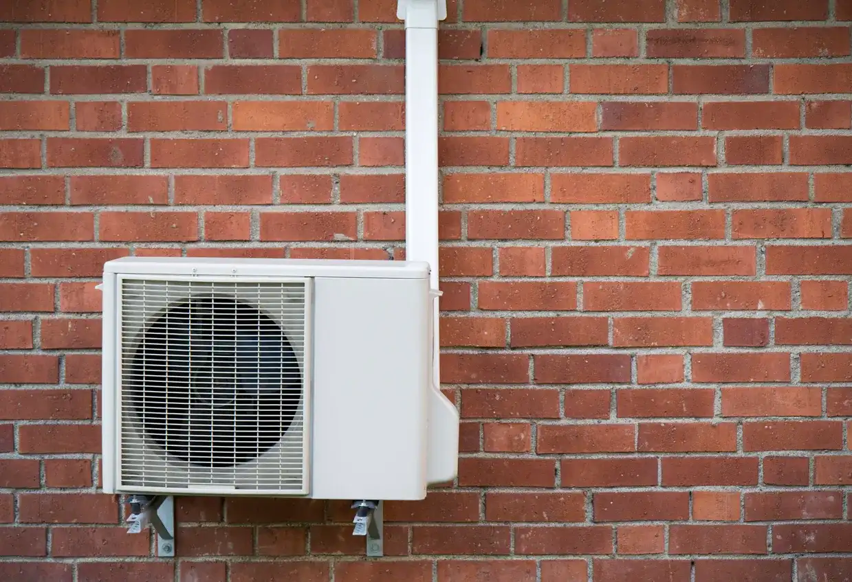 A white, wall-mounted heat pump unit is installed on a red brick wall, with a white pipe running up the side.