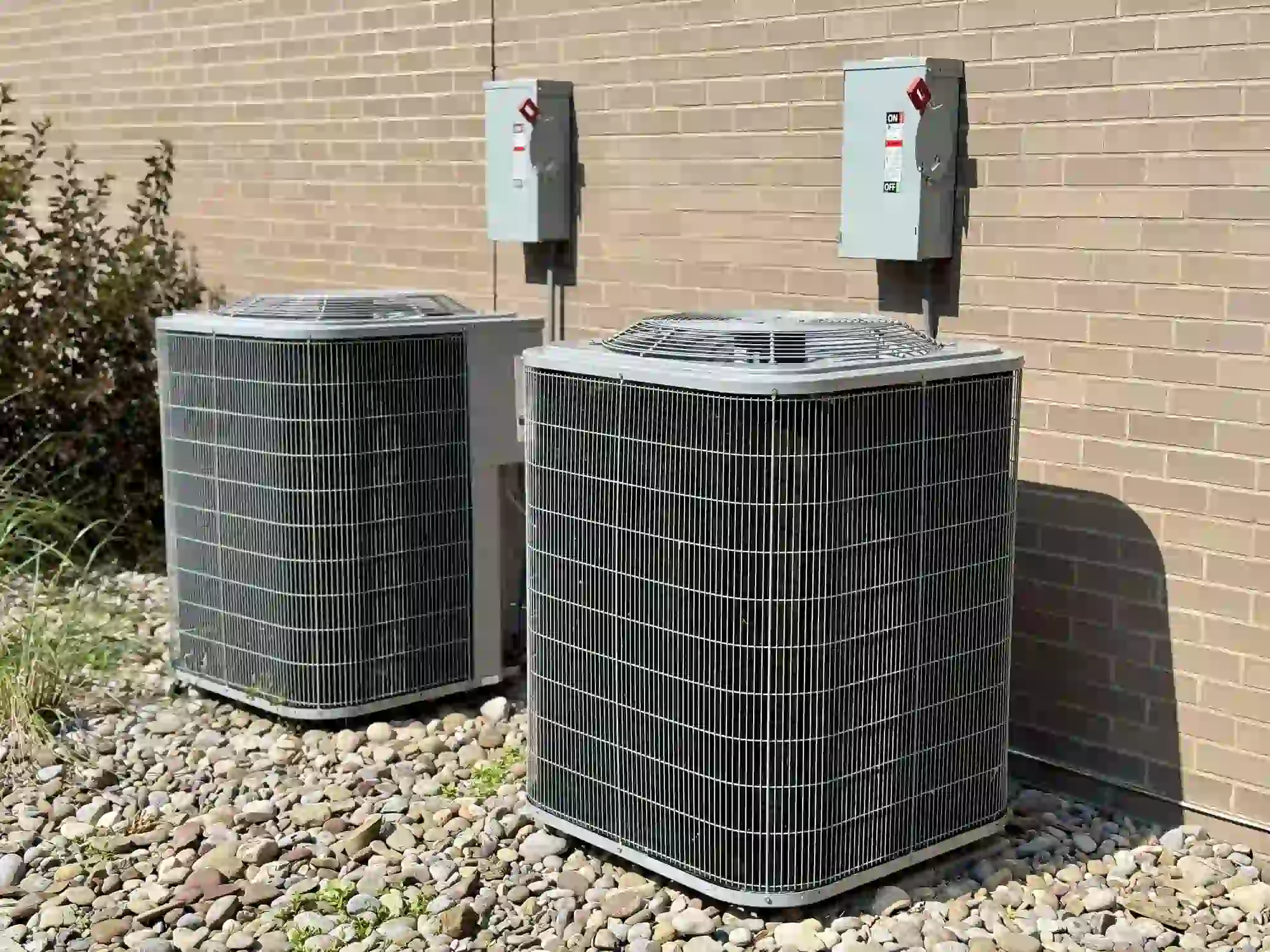 Two grey central air conditioning units or heat pumps sit on a bed of river rocks next to a tan brick wall. Each unit is connected to an electrical disconnect box mounted above.