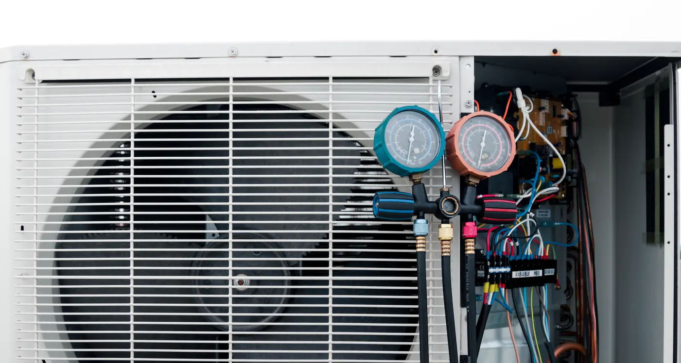 A close-up view of a manifold gauge set with teal and orange dials hanging on a white outdoor heat pump unit. The internal electrical wiring and a large fan are visible behind the grille.