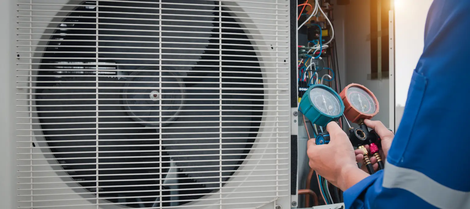 A technician in a blue uniform uses a manifold gauge set with teal and orange dials to test an outdoor heat pump unit. The unit's internal wiring and fan are visible behind the grille.