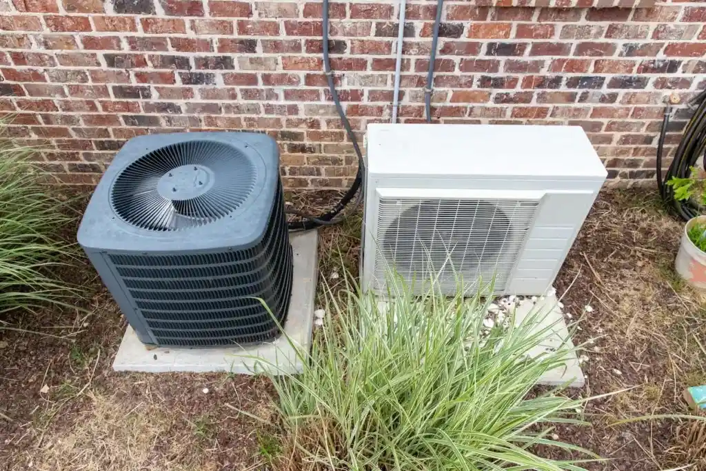 Two outdoor air conditioning condenser units sit side-by-side on concrete pads against a brick wall. A standard square unit is on the left, and a rectangular mini-split unit is on the right, surrounded by landscaping.