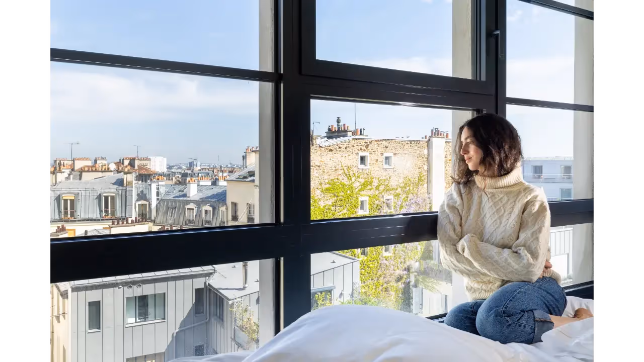 Woman in bed looking out a window in an apartment hotel furnished for a long term stay, Yuna.