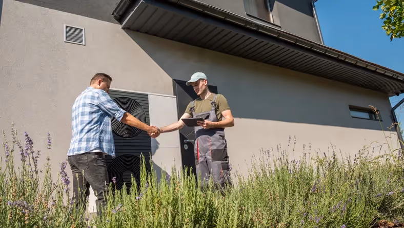 A man in a plaid shirt is shaking hands with a technician in a gray uniform and blue hat in front of a modern ac unit mounted on the side of a gray house.