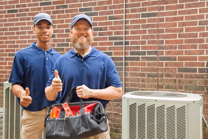 Two technicians pose in front of an air conditioning unit, one giving a thumbs up while holding a tool bag filled with equipment.