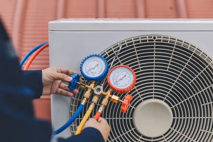 A technician adjusts blue and red pressure gauges on an air conditioning unit, surrounded by colorful hoses against a red rooftop backdrop.