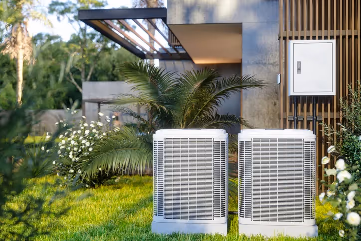  Two identical outdoor AC compressor units are placed side-by-side in a well-manicured green lawn with white flowers and palm plants in front of a modern home with wood paneling.