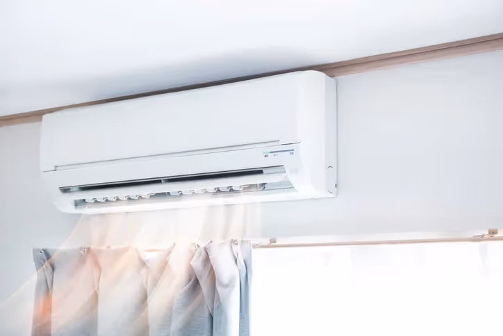A modern white air conditioning unit mounted on a wall, next to a window with blinds.