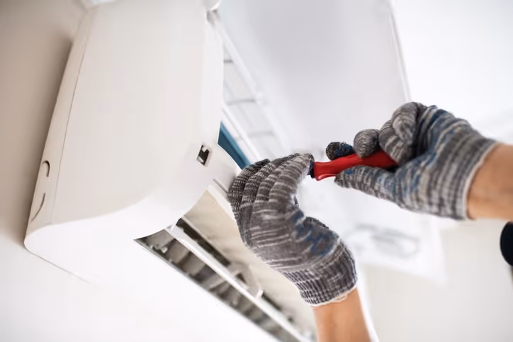 A person wearing gray gloves uses a red-handled screwdriver to repair a white wall-mounted air conditioner