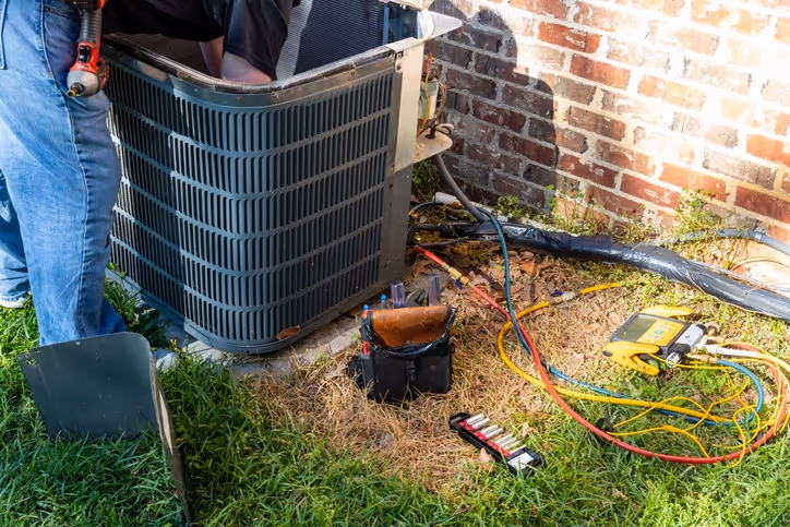 A technician repairing an outdoor air conditioning unit beside a brick wall.