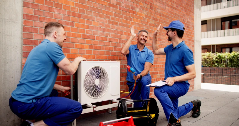 HVAC technicians in blue uniforms high-five joyfully beside an outdoor air conditioning unit on a brick wall.