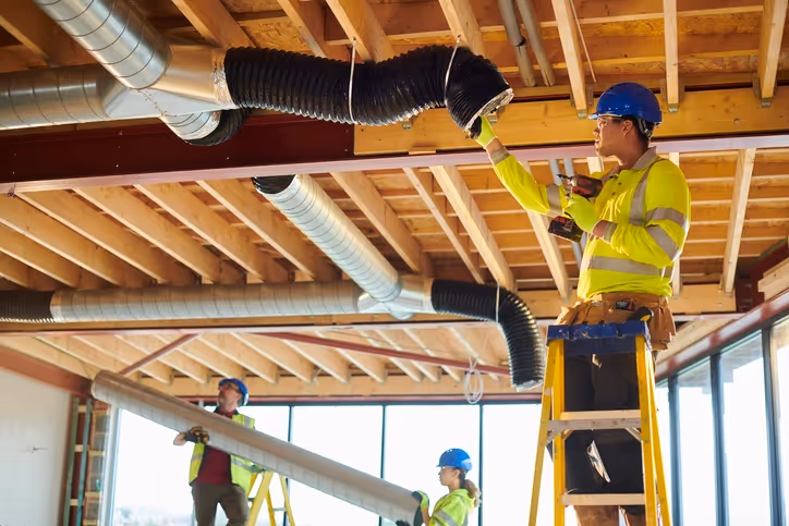 Three construction workers in safety gear and blue helmets install ductwork in a building with exposed wooden beams. 